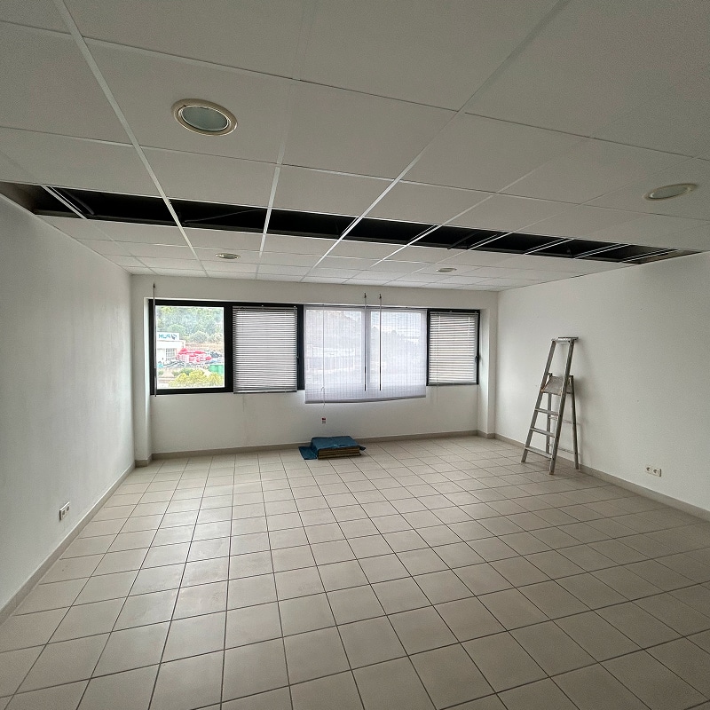 Bureau vide avec sol carrelé, murs blancs, fausses-fenêtres à stores et une ouverture rectangulaire dans le faux-plafond. Un escabeau est près de la fenêtre.