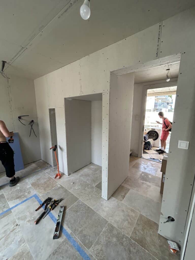 Chantier rénovation intérieure : pose de plaques de plâtre (BA13, inscription "Recyclée et recyclable avec E"), outils, carreaux de travertin et ouvrier au travail.