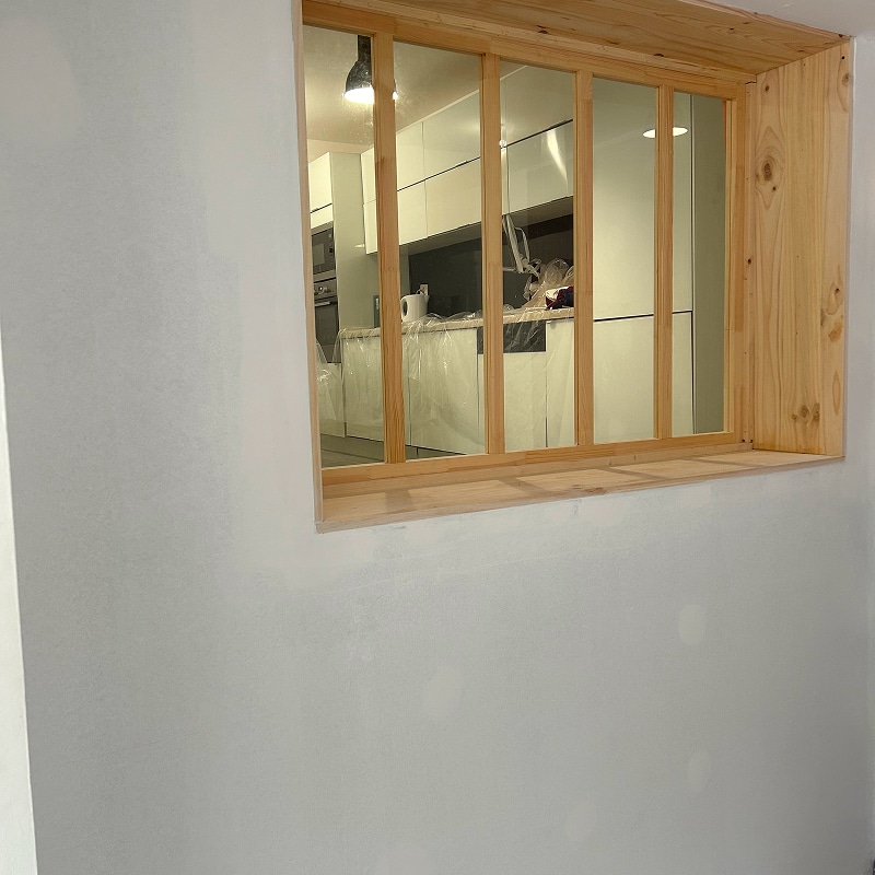 Passer-plat en bois clair avec vitrage quadrillé, donnant sur une cuisine moderne blanche en cours de rénovation, murs gris clair.
