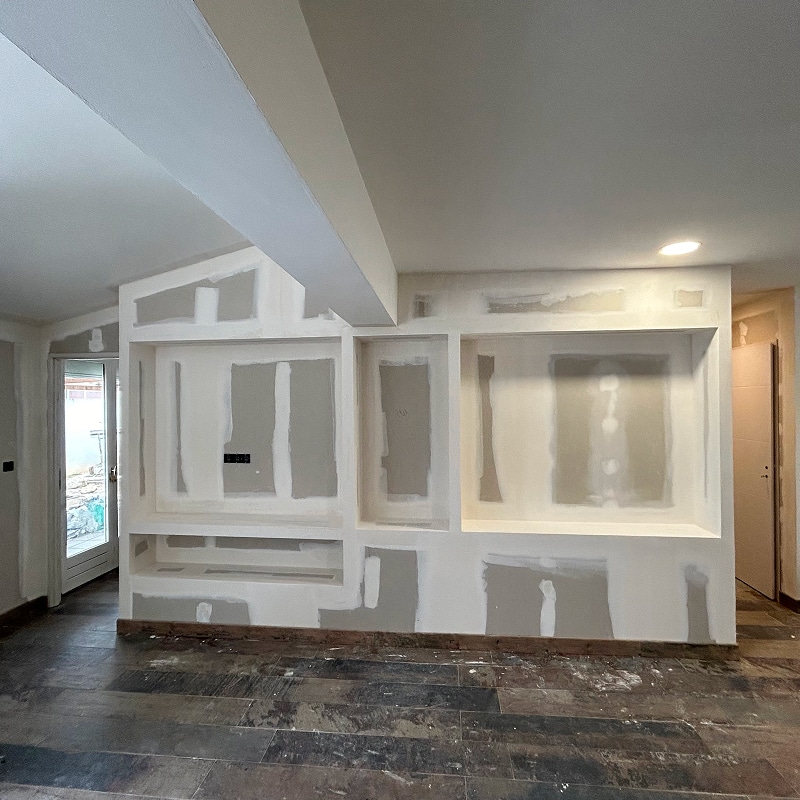 Mur de plaques de plâtre (gypse) en cours de jointoiement, avec niches intégrées pour meuble TV, sol sombre et porte vitrée menant à l'extérieur.