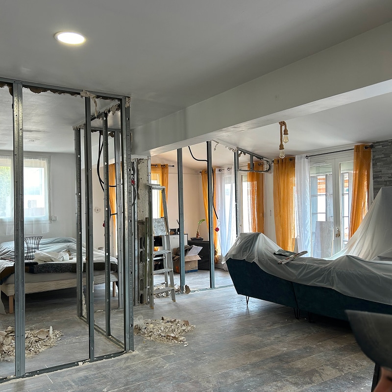Chantier de rénovation : structures métalliques de cloisonnement en cours dans un salon avec canapé bâché et rideaux orange.