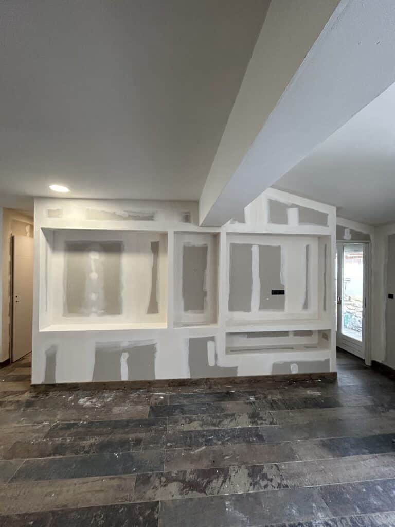 Mur en plaques de plâtre fraîchement jointoyées pour meuble TV ou bibliothèque, sol sombre en lames, lumière encastrée.