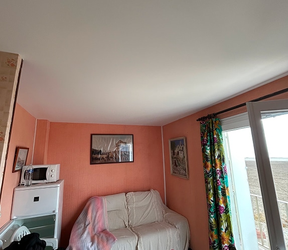 Intérieur d'une pièce aux murs saumon clair, avec un canapé clair, un petit frigo et micro-ondes, et une porte-fenêtre donnant sur un extérieur plat et clair.