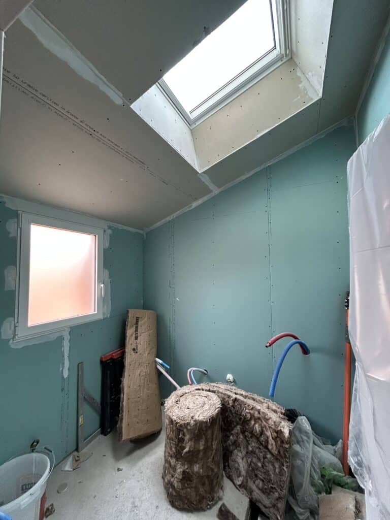 Plaques de plâtre, chantier et isolation en cours Chantier intérieur : murs et plafond en plaques de plâtre (vertes et blanches) avec joints enduits. Fenêtre et Velux laissent entrer la lumière. Isolation KNAUF en rouleaux sur le sol.