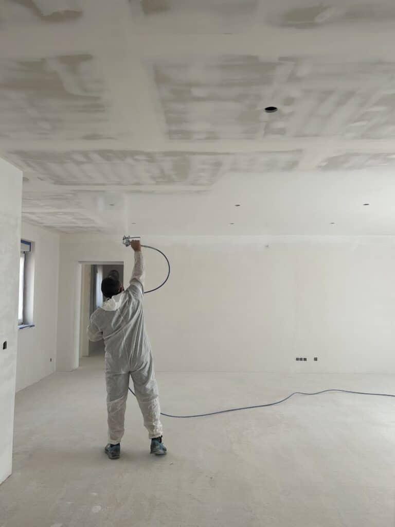 Application d'enduit au pistolet sur plafond de placo Peintre en combinaison de protection pulvérise un enduit de finition sur un plafond de plaques de plâtre brut dans une pièce en construction.