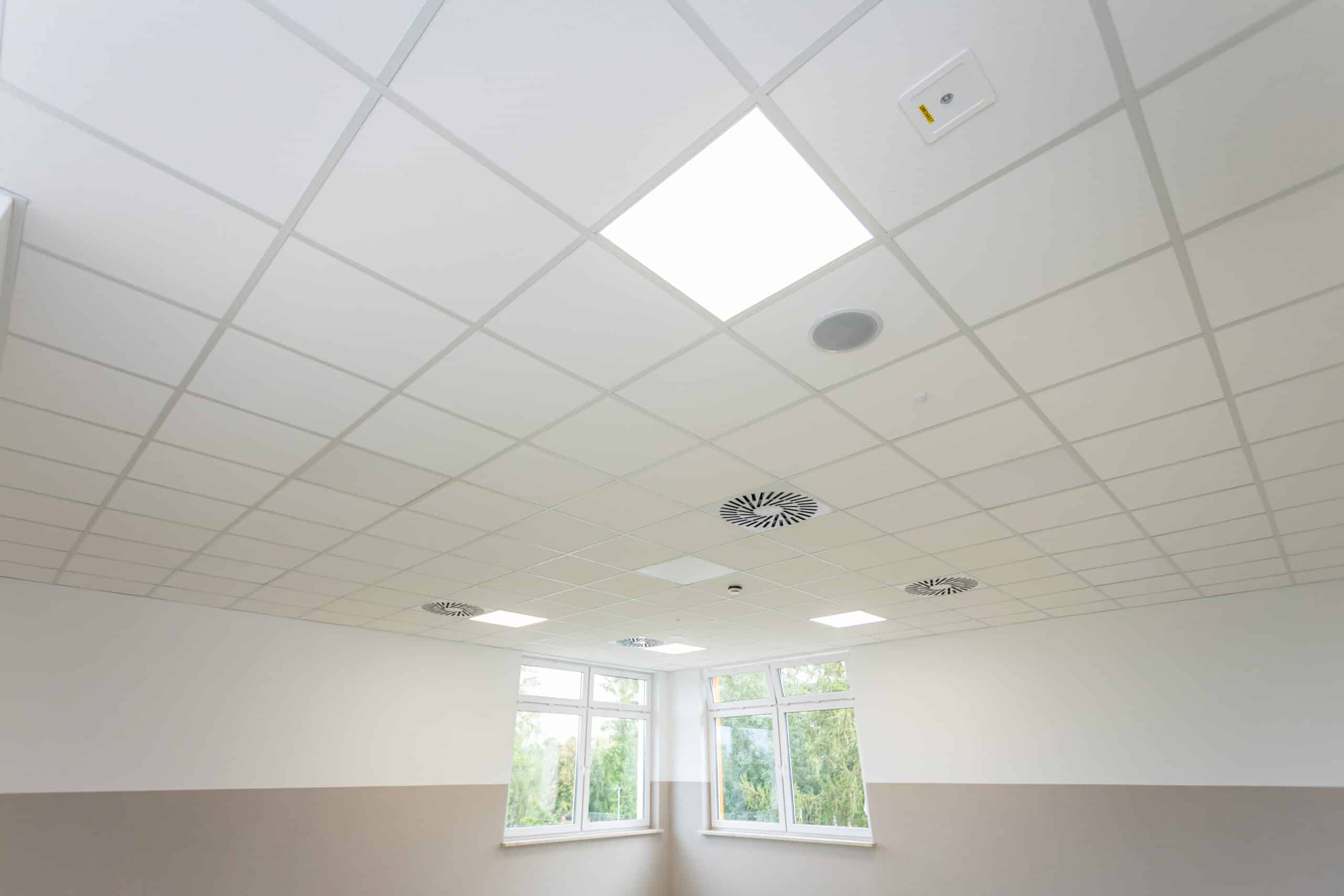 Plafond blanc à dalles modulaires avec éclairage encastré, diffuseurs d'air circulaires, et un appareil de sécurité étiqueté '10826057'.