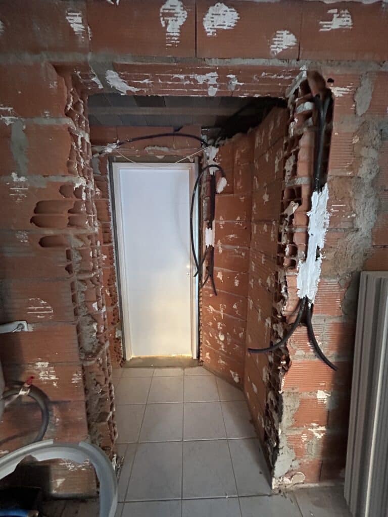 Chantier : Embrasure de porte en briques et câbles électriques Porte entrouverte dans une embrasure défoncée en briques creuses, câbles électriques noirs apparents et scellés au plâtre blanc, chantier en cours.