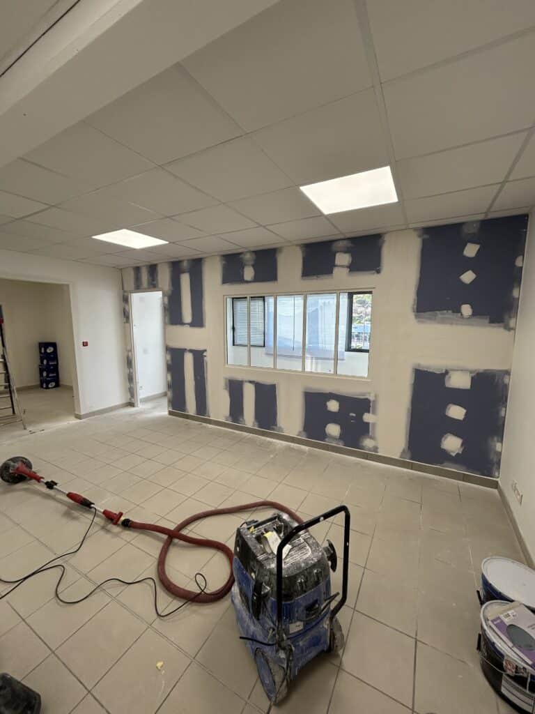 Rénovation murs placo : ponçage et jointoiement en cours Ponçage de murs en plaques de plâtre après jointoiement, avec aspirateur industriel bleu et ponceuse girafe rouge dans un bureau en rénovation.