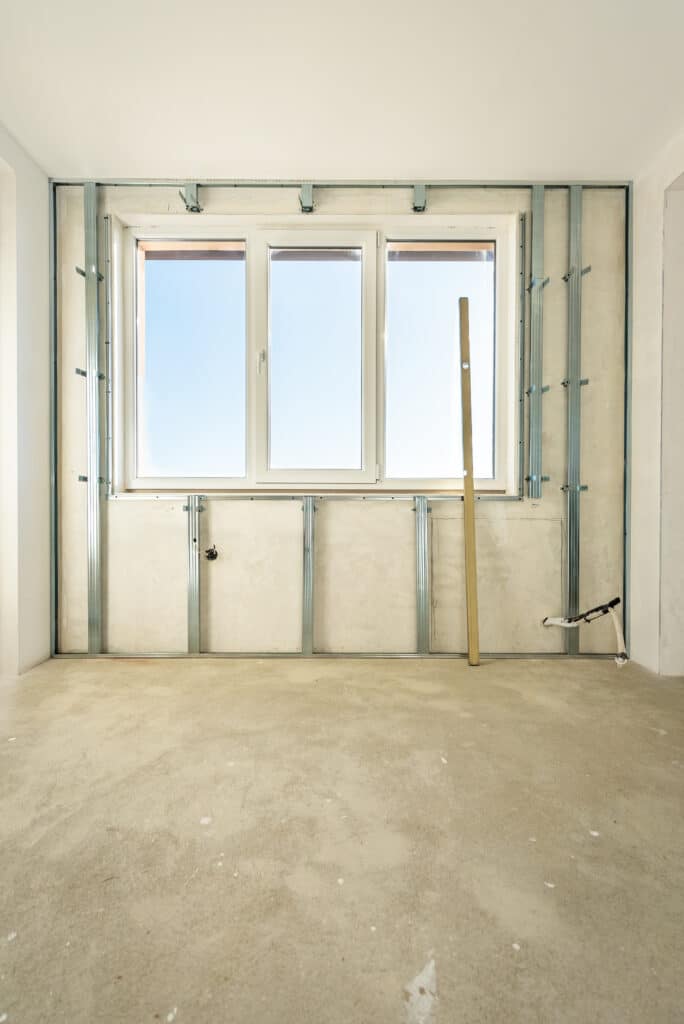 Pose de plaques de plâtre : Ossature métallique près fenêtre Structure métallique pour cloison sèche devant une fenêtre PVC triple, sol en béton brut et niveau à bulle en bois pendant la rénovation.