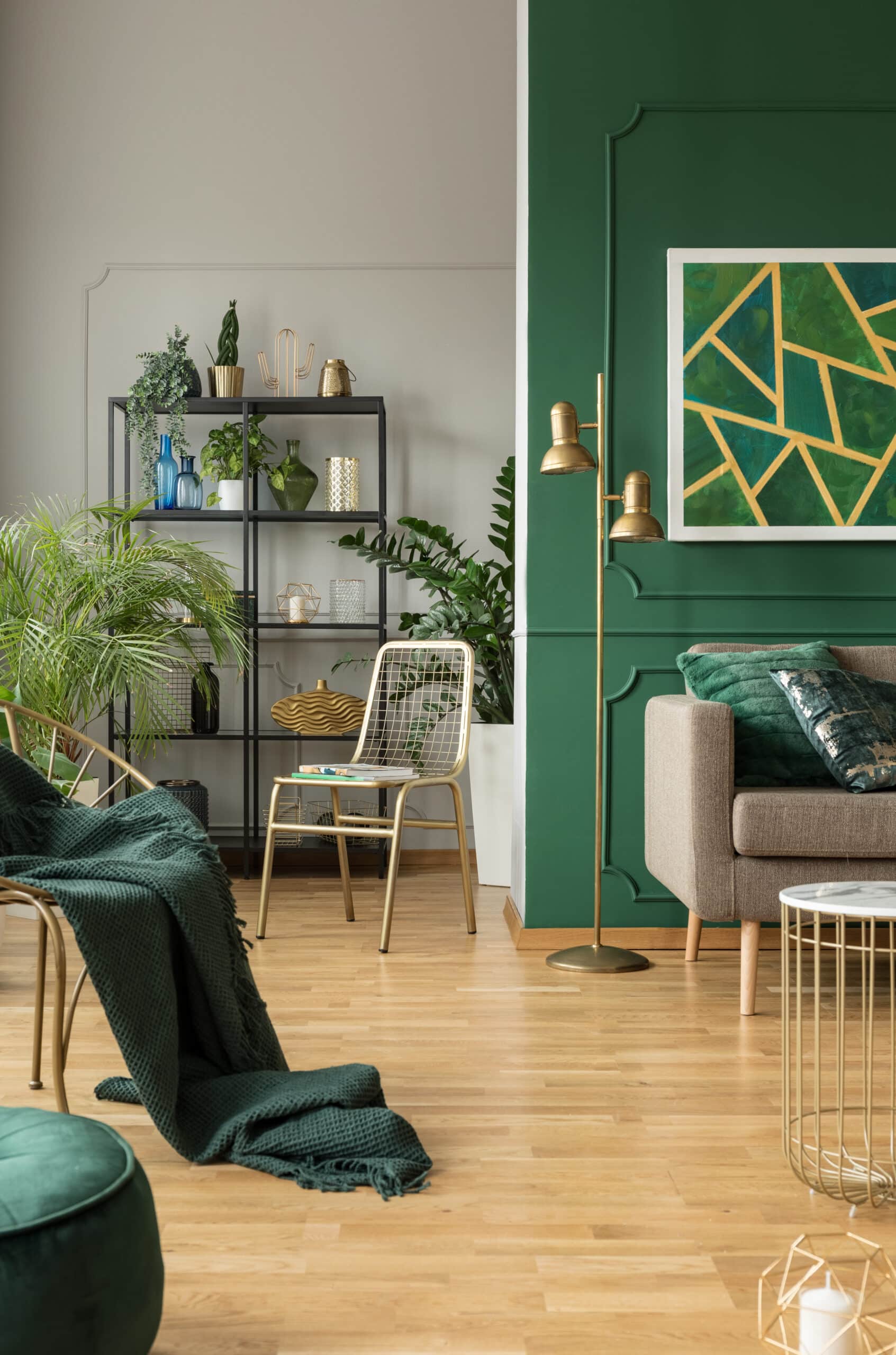 Salon moderne avec murs gris et vert émeraude, étagère noire remplie de plantes, chaise dorée et canapé beige.
